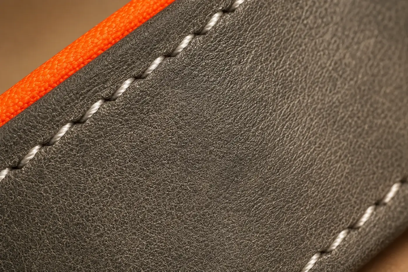 Extreme macro of the gray leather strap showing its grain, two rows of white contrast stitching and the orange lining along the edge